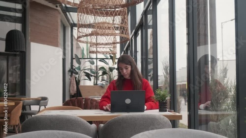 A beautiful young girl in a red jacket is sitting at a table in a restaurant. Working at a laptop. Remote work concept.