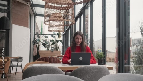 A beautiful young girl in a red jacket is sitting at a table in a restaurant. Working at a laptop. Remote work concept.