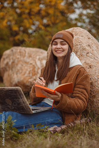 Portrait of smiling freelancer with laptop writing in notepad in park and looking at camera 