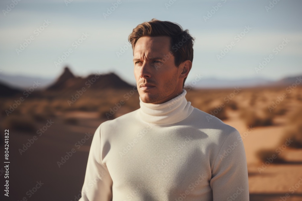 Portrait of a blissful man in his 30s wearing a classic turtleneck sweater while standing against backdrop of desert dunes
