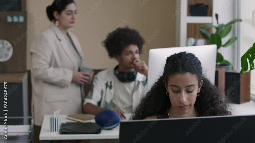 Young Biracial female programmer working on desktop computer at desk in modern IT office while two diverse colleagues looking at desktop monitor in background Young Biracial female programmer working on desktop computer at desk in modern IT office while two diverse colleagues looking at desktop monitor in background