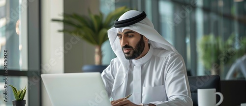 Working on Laptop Computer in an Office. Successful Arab Businessman in White Traditional Outfit. Saudi, Emirati, Arab Businessman Concept. photo