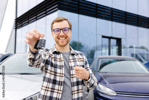 Emotional guy holding key in hand and smiling at camera. Man showing key from his new electric car. Young man buying auto at dealership salon