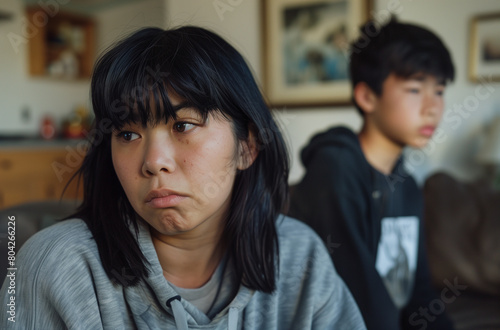 Asian woman with black hair and bangs sitting on the couch in her living room, worried about her teenage son standing behind her.