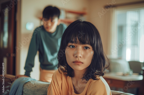 Asian woman with black hair and bangs sitting on the couch in her living room, worried about her husband standing behind her. relationship problems and family tension.