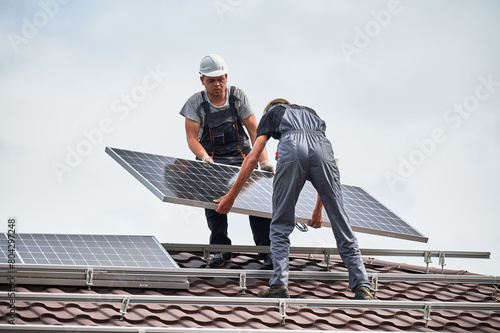 Men technicians carrying photovoltaic solar modul on roof of house. Workmen in helmets installing solar panel system outdoors. Concept of alternative and renewable energy.