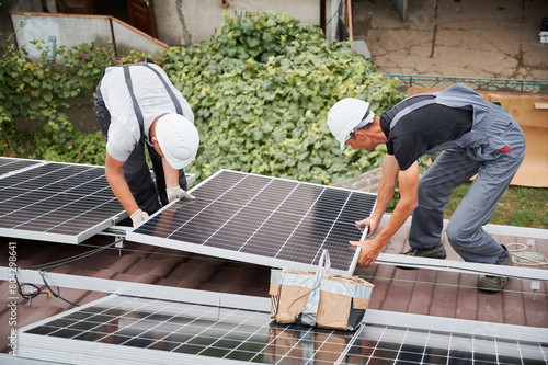 Men technicians installing photovoltaic solar moduls on roof of house. Engineers in helmets building solar panel system outdoors. Concept of alternative and renewable energy.