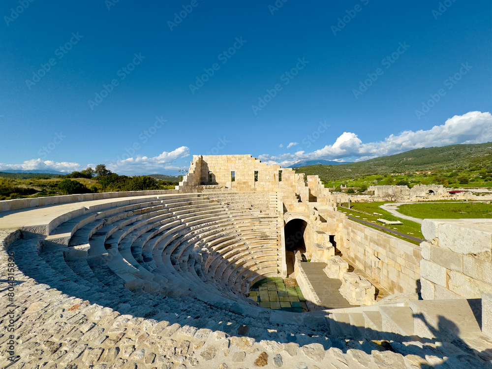 The ancient amphitheater of Patara in Antalya, Turkey, features stone seating and arches, set against a scenic backdrop. A stunning example of historical architecture and cultural heritage.