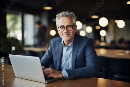 Portrait of a smiling senior businessman working on his laptop in a modern office, wearing a blue suit jacket with glasses, sitting behind a wooden table.