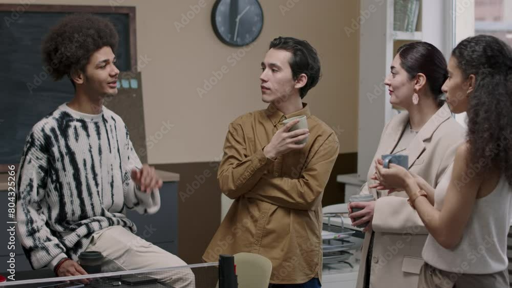 Medium shot of four young diverse office employees standing next to tennis table at workplace and having conversation during coffee break Medium shot of four young diverse office employees standing next to tennis table at workplace and having conversation during coffee break