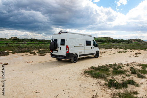 Solitary Camper Van Journey. A white Camper van parked on a sandy path near grassy hills under a cloudy sky. Parked in a desert.