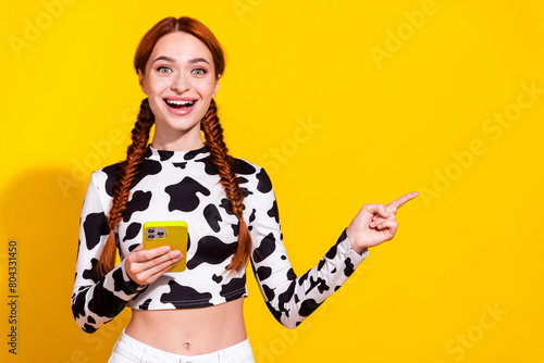 Photo of impressed funky woman wear cow skin print top texting messages gadget pointing empty space isolated yellow color background