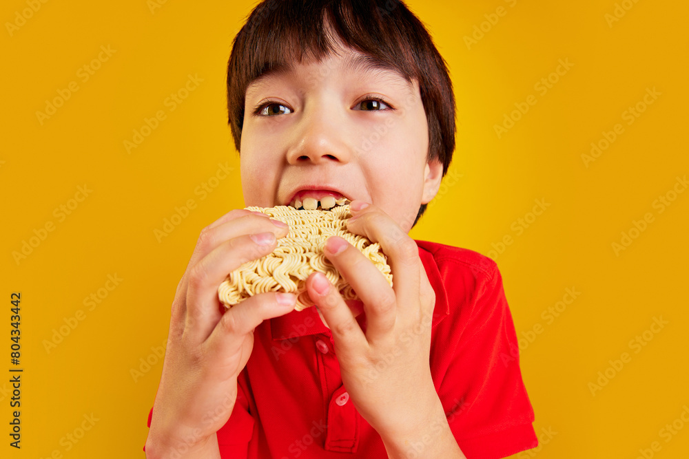 Young boy, child in a red polo shirt enjoying noodles, eating with his hands against yellow background. Concept of food, childhood, emotions, meal, menu, pop art. Close-up portrait Young boy, child in a red polo shirt enjoying noodles, eating with his hands against yellow background. Concept of food, childhood, emotions, meal, menu, pop art. Close-up portrait
