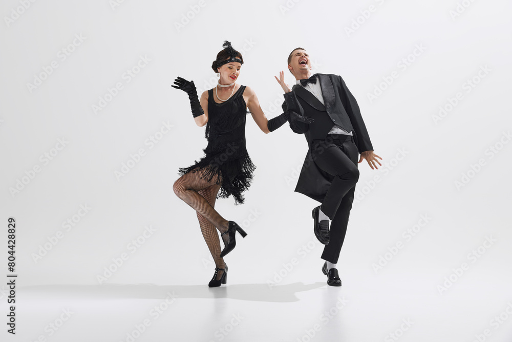 Obraz premium Woman in black flapper dress dancing with man in classic black suit, showcasing lively spirit of the 1920s isolated over white studio background. Concept of art, retro and vintage, entertainment, 20s