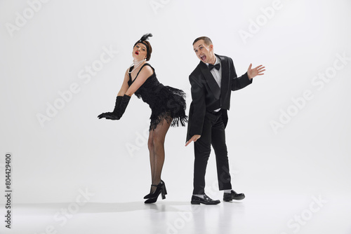Obraz na plátně Playful dance scene featuring couple dressed in 1920s fashion, stylish young man and woman dancing isolated over white studio background