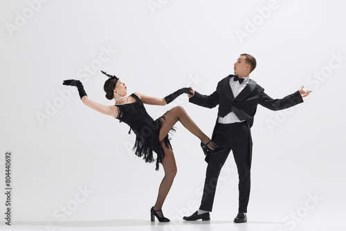 Photography Stylish couple reenacts 1920s dance, woman in fringe dress and man in dapper black suit dancing isolated over white studio background