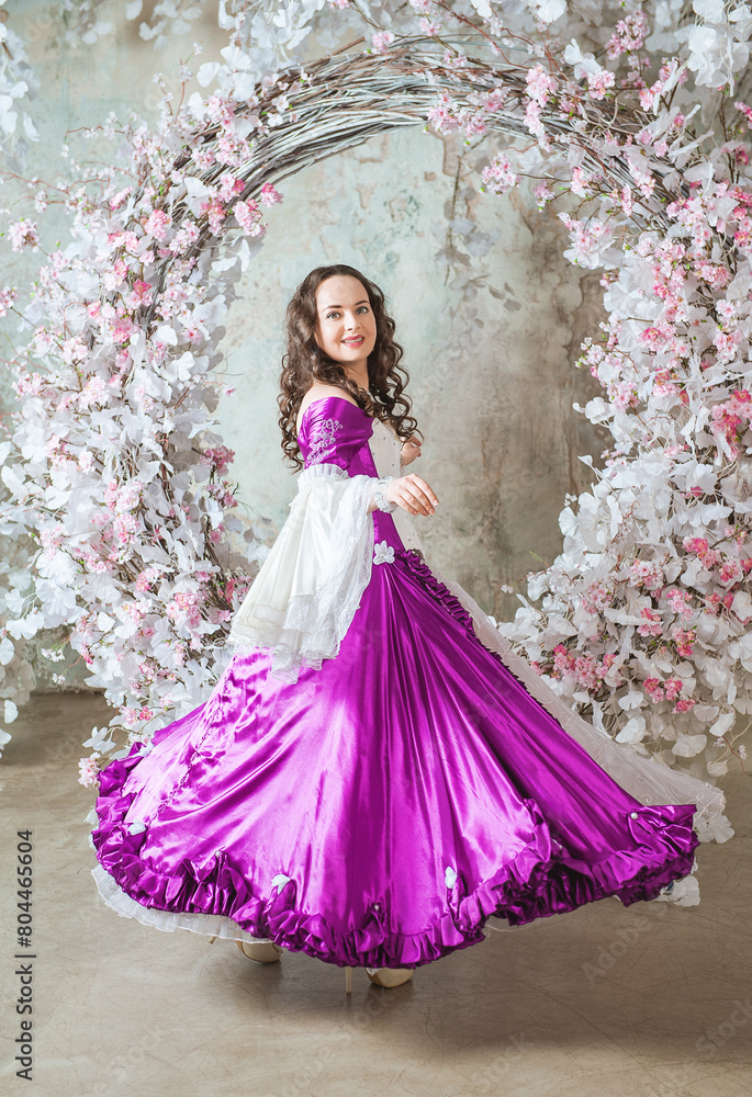 Fototapeta premium Happy beautiful woman in fantasy white and purple rococo style medieval dress dancing near decoration with white flowers