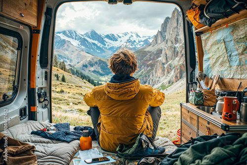 A young man sitting in his camper looks out at the mountain panorama from the open rear door, seen from behind
