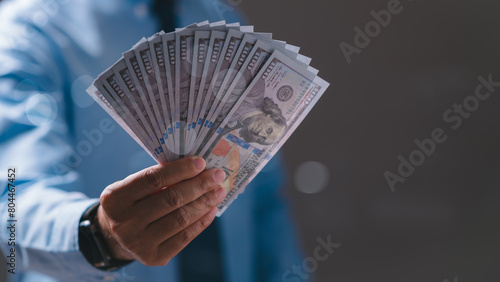 A man is holding a bunch of money in his hand. The money is in the form of a stack of one hundred dollar bills. The man is wearing a blue shirt and a tie. Concept of wealth and prosperity