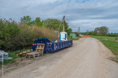 Next to a road is a large blue container for construction waste and a construction site toilet