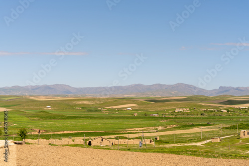 A large  open field with a few houses in the distance. The sky is clear and blue