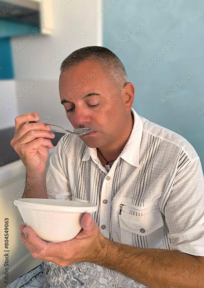 Young handsome adult man,sitting in the kitchen at home,with a bowl in his hand and a spoon eating.Eating healthy breakfast at home.Healthy afternoon snack. Young handsome adult man,sitting in the kitchen at home,with a bowl in his hand and a spoon eating.Eating healthy breakfast at home.Healthy afternoon snack.