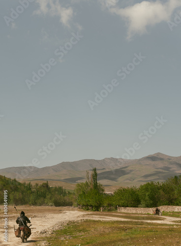 A man rides a motorcycle down a dirt road in a desert. The sky is clear and the sun is shining brightly