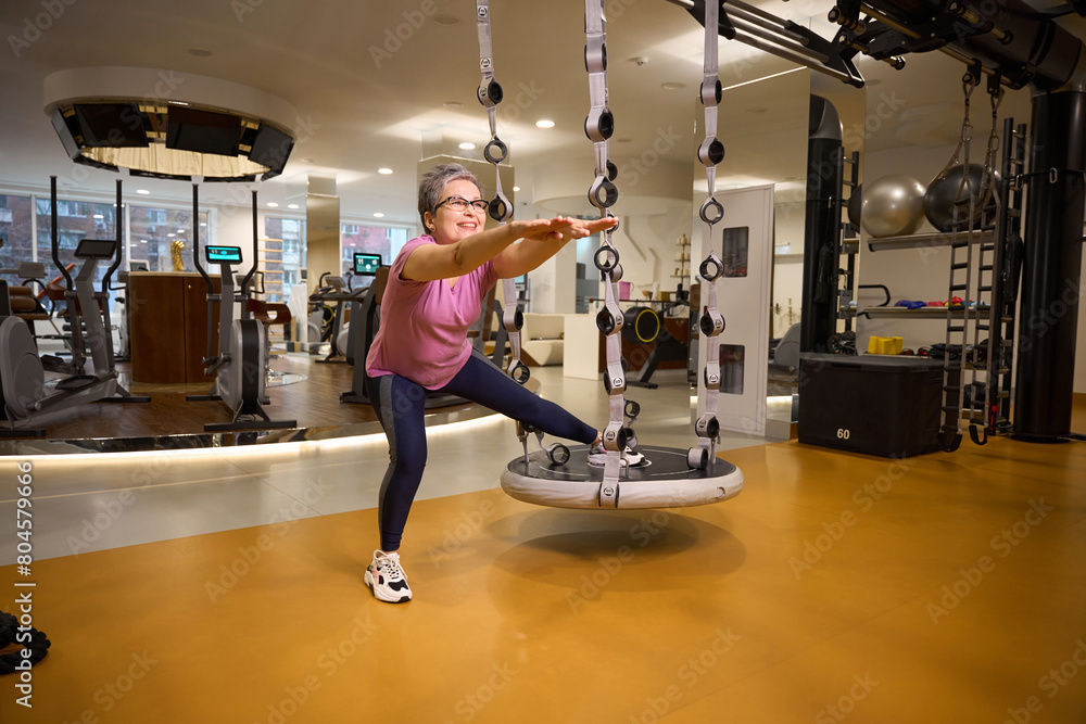 Middle aged smiling caucasian woman doing sport exercise on hanging mat in gym