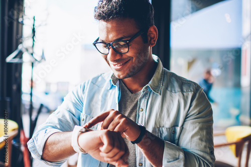 Foto Cropped image of good looking smiling hindu businessman in cool spectacles configuring digital smartwatch sitting indoors