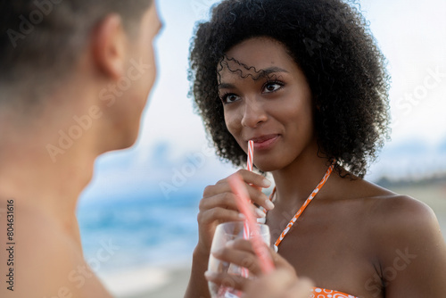 Close-up of a young woman - smiling and sipping a cocktail while looking at her partner, against a beach sunset backdrop - romantic, relaxed. (ID: 804631616)