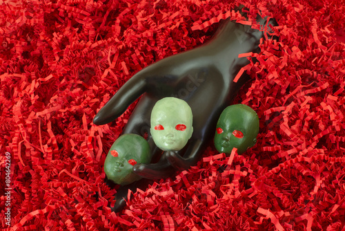 Three spooky creepy green Martian heads with red eyes with black mannequin hand emerging from red shredded paper background