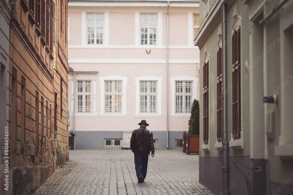 Man walking down an old street, view from the back. Rear view of a stylish man in a hat and coat walking down the street.