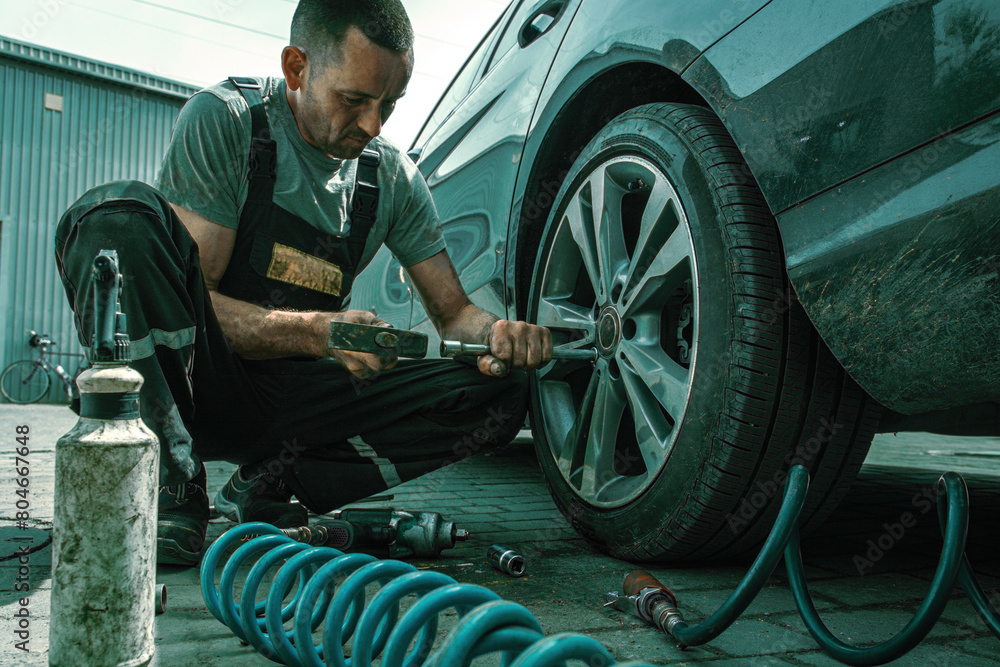 A car service technician in overalls repairs a wheel and knocks out the wheel nuts with a hammer. A car service technician in overalls repairs a wheel and knocks out the wheel nuts with a hammer.