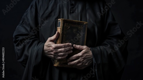 Catholic christian church priest wearing black cassock robe holding the holy bible book in his hands. Isolated on dark black background photo