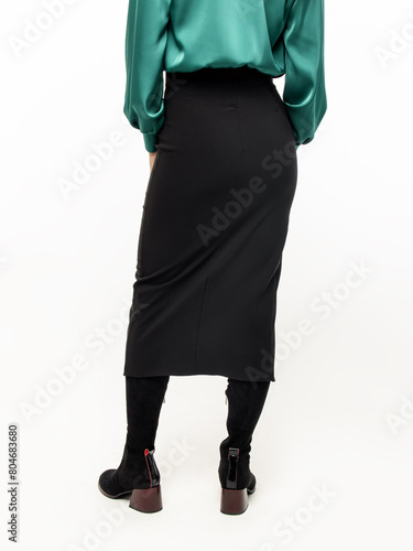 Woman in black straight midi skirt with a slit on white background. Silk green blouse and high boots. Rear view
