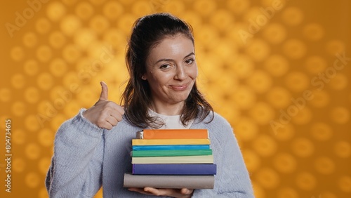 Portrait of happy woman with stack of books in hands showing thumbs up, studio background. Joyous bookworm holding pile of novels, feeling upbeat, doing positive hand gesturing, camera B photo