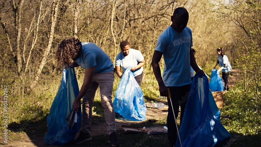 Fototapeta premium Diverse men volunteers pick up rubbish and plastic trash with tongs, working to combat illegal dumping and preserve natural forest environment. Activists volunteering for litter cleanup. Camera B.