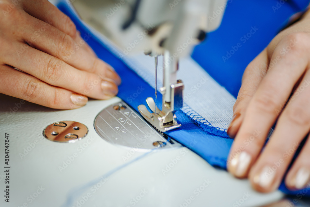 sewing machine with needle and thread on table in atelier, close-up view of hands of working sewer, blue fabric and needle, handmade and sewing concept