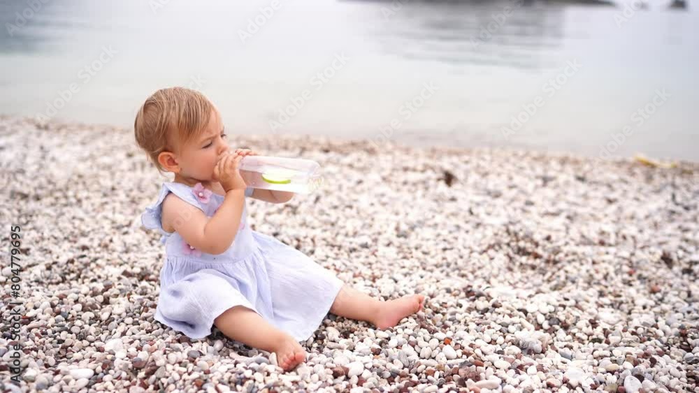 Little girl drinks water from a bottle while sitting on a pebble beach by the sea. High quality 4k footage Little girl drinks water from a bottle while sitting on a pebble beach by the sea. High quality 4k footage
