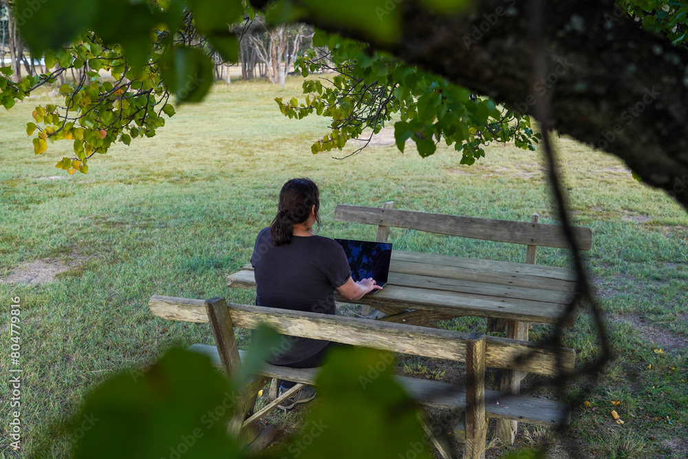Fototapeta premium Woman working remotely on a wooden bench under a tree
