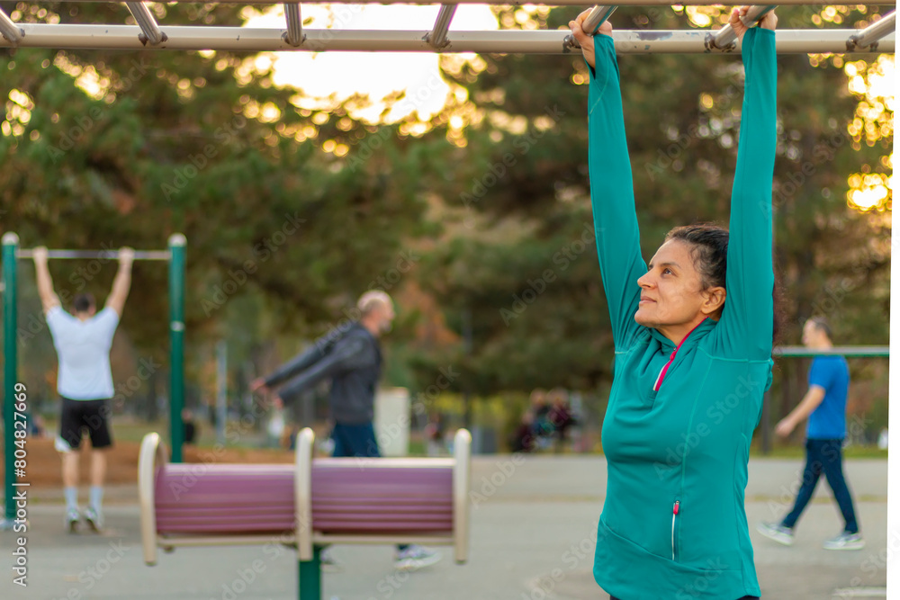 Fototapeta premium Front view of mature woman wearing sports clothes doing pull-ups during a workout on a promenade. Healthy, sporty lifestyles and slimming concept