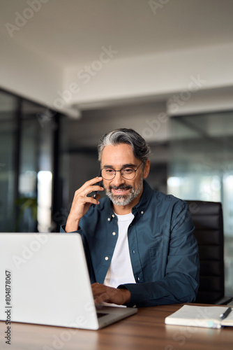 Happy middle aged business man talking on cell phone working on laptop in office. Busy mature professional businessman executive making call on cellphone using computer sitting at desk. Vertical.