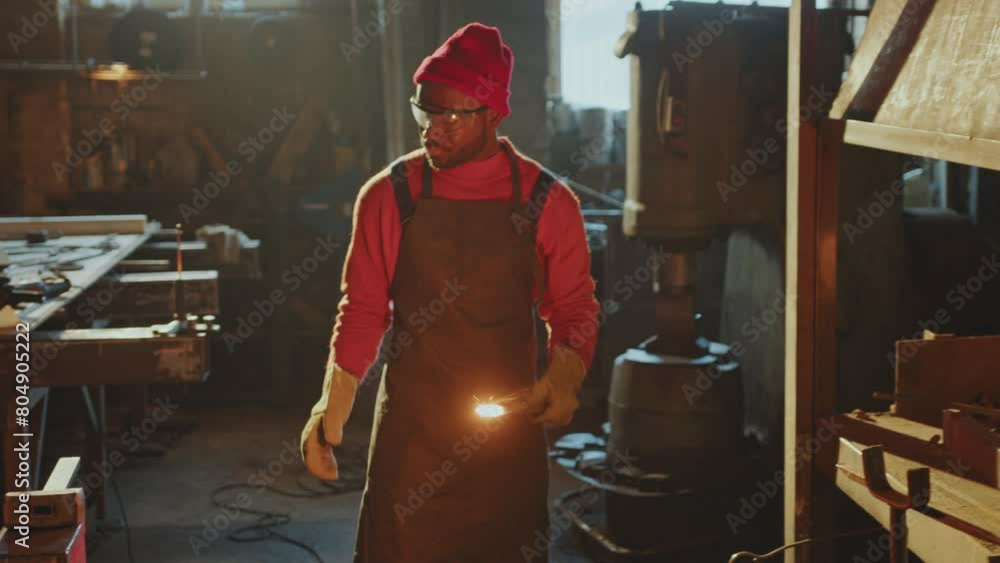 African American blacksmith in safety glasses, gloves and apron removing red-hot metal workpiece from fire in charcoal forge and shaping it with sledgehammer on anvil in the workshop African American blacksmith in safety glasses, gloves and apron removing red-hot metal workpiece from fire in charcoal forge and shaping it with sledgehammer on anvil in the workshop