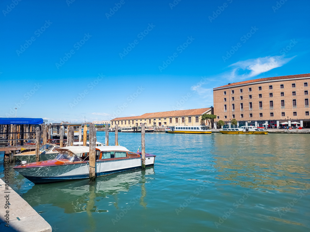 The boat peacefully rests in a serene harbor next to a majestic building, with a tranquil canal nearby, reminiscent of Venice. The clear blue sky reflects on the waters, creating a picturesque scene.