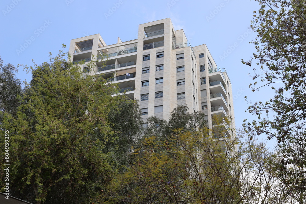 Buildings and structures in Tel Aviv against the background of branches and leaves of tall trees.