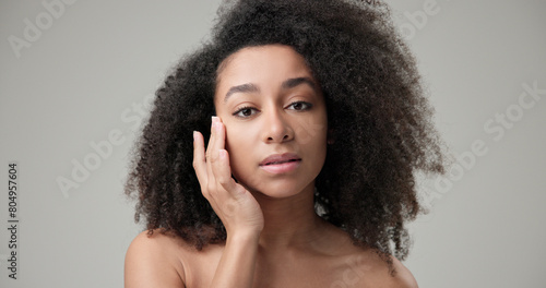 Beauty and healthcare concept - beautiful African American woman with curly afro hairstyle and clean, healthy skin touches her cheek and face with her hand, posing and looking at the camera © Anatoly Repin