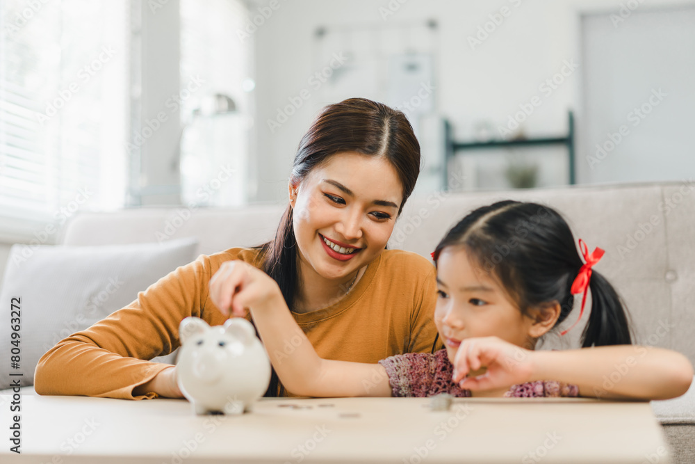 Cute little asian girl saving money by adding a coin in piggy bank with mother at home Cute little asian girl saving money by adding a coin in piggy bank with mother at home