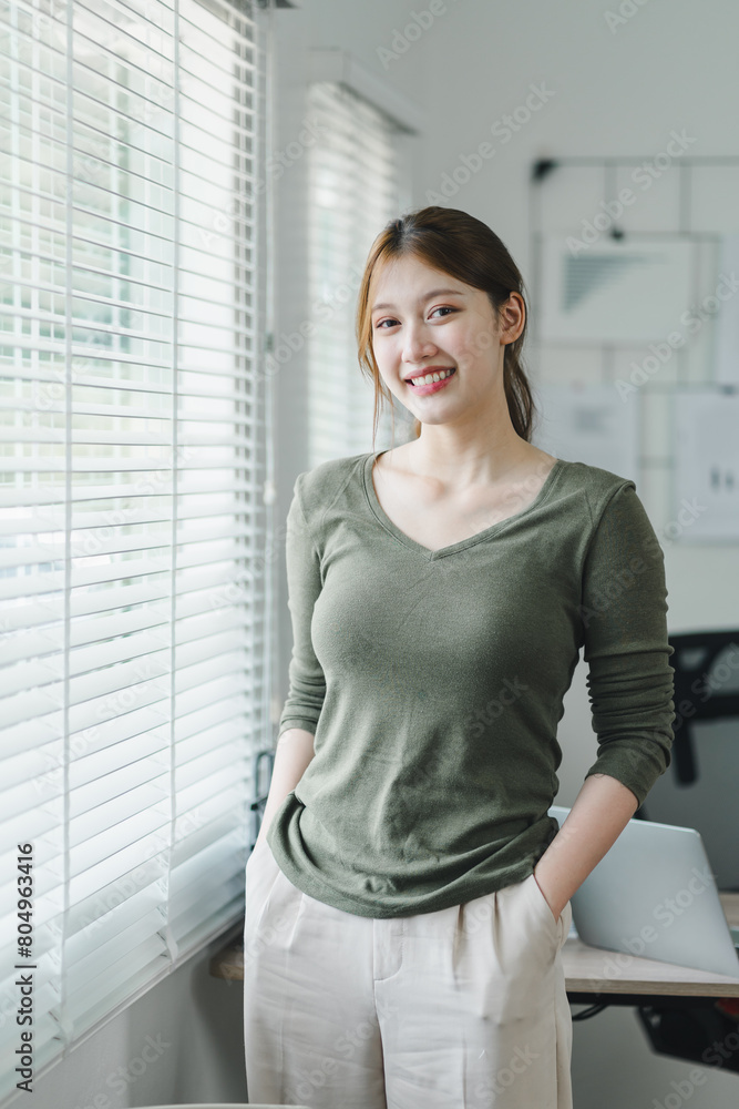 Smiling asian young businesswoman standing by the window in a bright office environment. Smiling asian young businesswoman standing by the window in a bright office environment.
