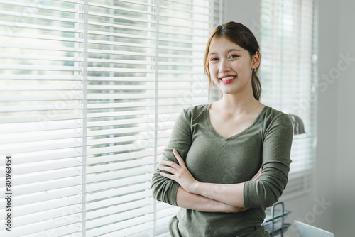Smiling asian young businesswoman standing by the window in a bright office environment.