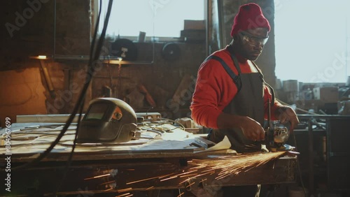 African American blacksmith in apron and safety glasses cutting ferrous metal with angle grinder producing sparks during the day at his workshop photo